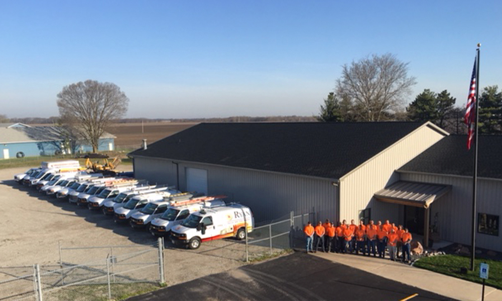 A wide view of the Ryan Electric parking lot showing a lineup of company service vans parked side by side. Employees wearing bright orange shirts stand together near the entrance of a beige metal building with a black roof. An American flag on a tall pole is visible to the right, and open fields and trees are in the background.