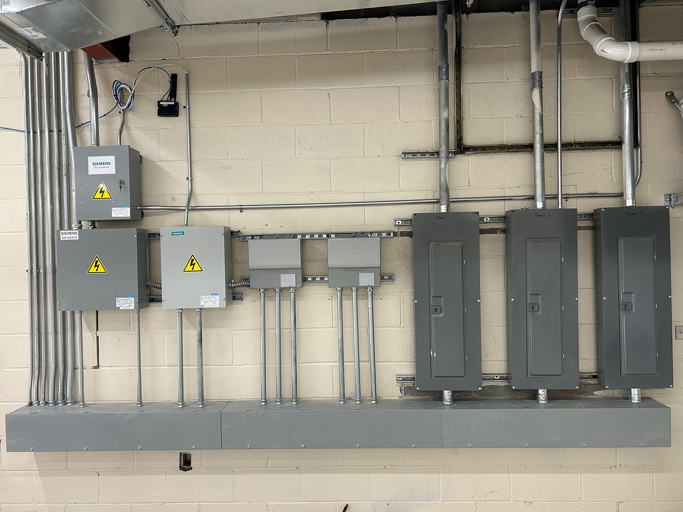 Commercial generator transfer switch and electrical panel installation featuring Siemens automatic transfer switches with yellow hazard labels, metal conduit runs, and multiple gray electrical distribution panels mounted on concrete block wall in industrial facility
